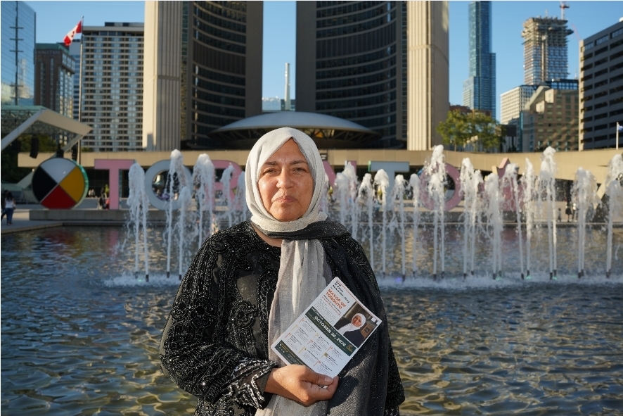 Bahira Abdulsalam at Nathan Phillips Square holding a campaign flyer, with City Hall and the TORONTO sign behind.