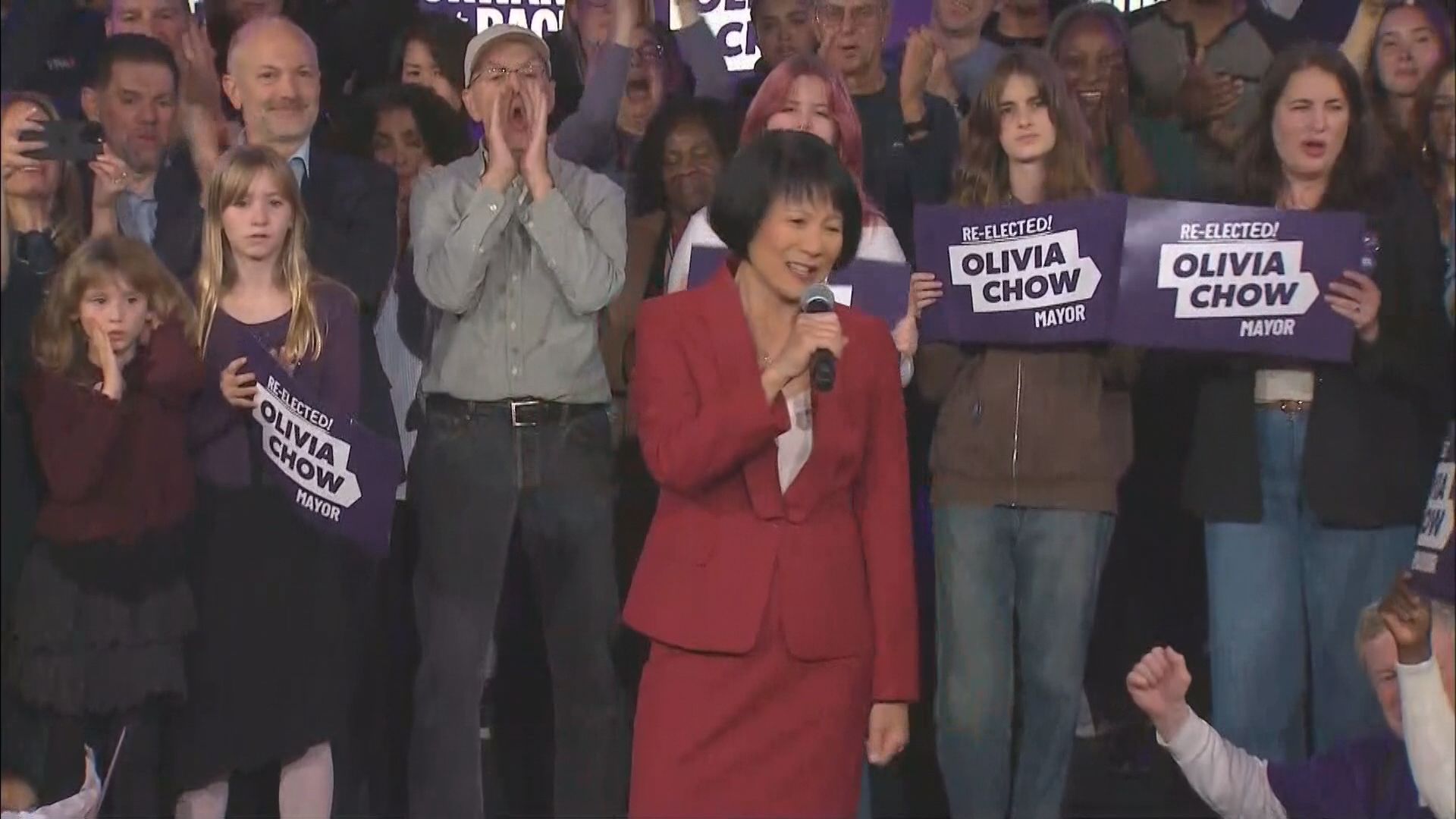 Olivia Chow speaking into a microphone at her election night victory rally surrounded by supporters holding Re-Elected! Olivia Chow Mayor signs