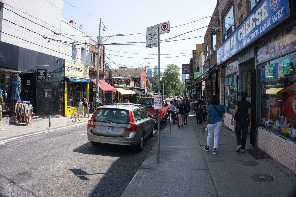 Looking south down Kensington Avenue in Toronto, with neighbourhood shopfronts and people walking on the sidewalk on a sunny day