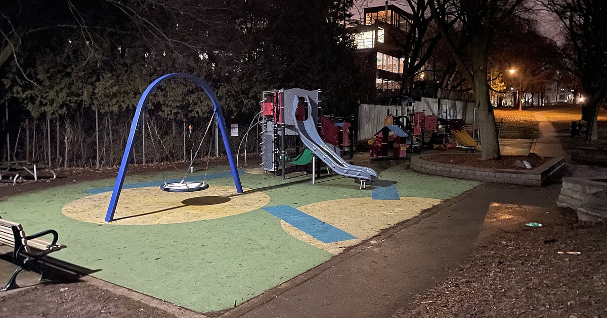An empty children's playground with swings and a slide in a Toronto park at night, lit by street lamps