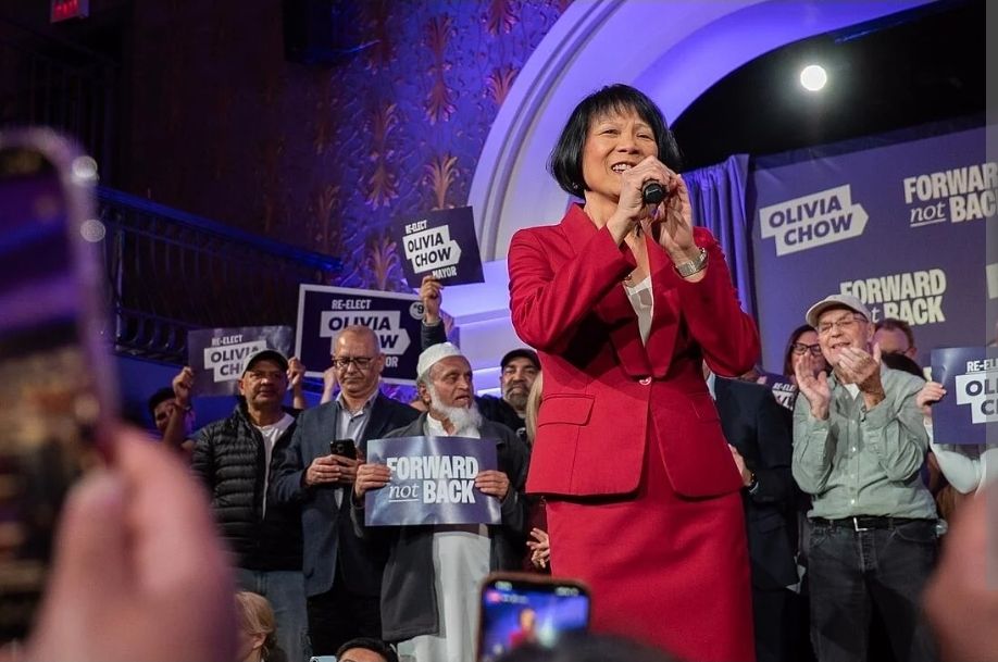 Olivia Chow speaking into a microphone at an election rally surrounded by supporters holding Re-Elect Olivia Chow Mayor and Forward Not Back signs