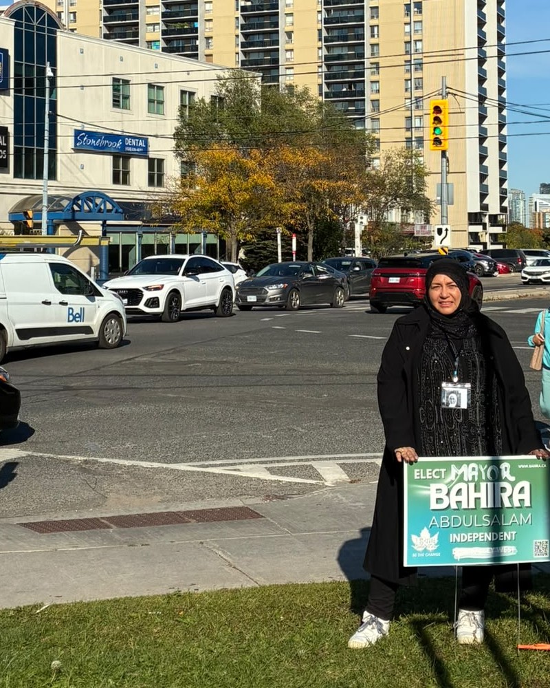 Bahira Abdulsalam stands on the grass at the Bathurst and Sheppard intersection holding her campaign lawn sign, with traffic and apartment towers behind her