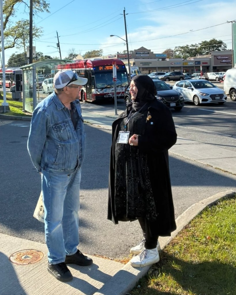 Bahira Abdulsalam talks with a resident in a denim jacket beside a TTC bus stop at Bathurst and Sheppard, with an 84 bus behind them
